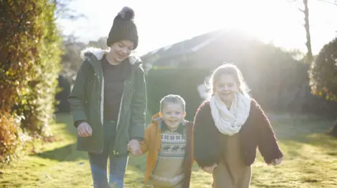 Getty Images Three children - two boys and a girl - smile and hold hands. All are dressed in warm clothes. They are outside on a sunny autumn day with golden leaves on a hedge and trees behind them.