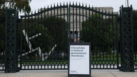 Getty Images A closed sign outside the National Gallery of Art in Washington, DC. The sign is in front of large, black, wrought iron gates behind which you can see a sculpture garden and a white, neo-classical building.