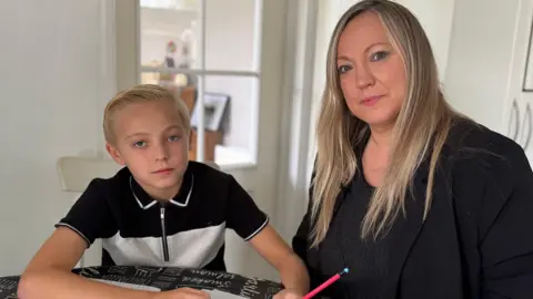 Charlie Rose/BBC Joshua, with short blonde hair, holds a pencil as he sits at the kitchen table with his mum, Nicky, who's dressed smartly in black and has long blonde hair. Both are looking at the camera.