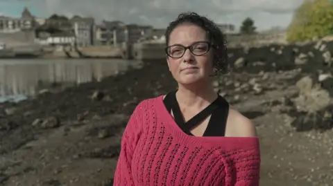 BBC Kelly Pardy stands in front of the sea. She is wearing a pink jumper and has black curly hair. She is also wearing black spectacles.
