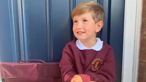 Family handout A young boy with dark blond hair is sitting on a step outside a blue door. He is wearing a burgundy school uniform with long grey socks and black shoes. The school uniform's jumper has a yellow circular logo, and he has a matching school bag next to him. He is looking up to the side and smiling.