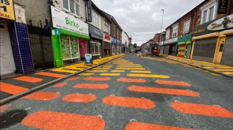 LDRS A street recently painted with unusual yellow and orange marking. Part of the walkway, which is flanked by shops, can be seen taped off, with painting vehicles seen parked up. 
