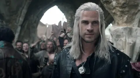 Netflix Liam Hemsworth as Geralt with black leather armour on and a sword holstered on his back. He has a silver pendant and long white hair. He stares into the distance. Behind him is a group of angry people in a medieval style area.