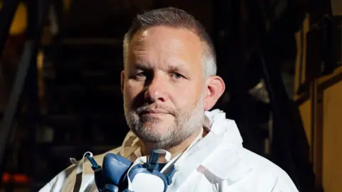 Owen Howells Photography Up close photograph of Ben Giles. His is aged 49, has grey short hair. He wears a  face mask around his neck. You can see his PPE around his neck, which is a hooded white suit. 