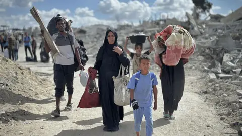 Anadolu via Getty Images A group of Palestinians walk on a road surrounded by rubble. A child leads the group, with two women behind him carrying belongings on their shoulder and arms. Behind them is a young girl carrying a saucepan on her head. To her right is a man with a large bag on his back. 