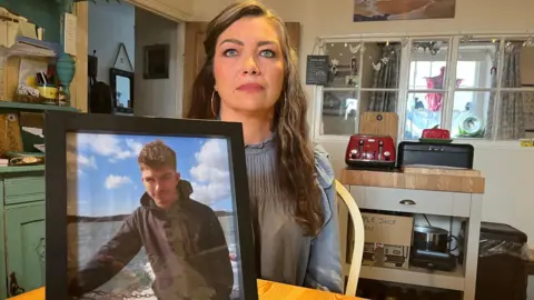 BBC A woman with long dark hair, wearing a blue top and sat in her home, is holding up a picture of a young man with dark hair and a black hoody 