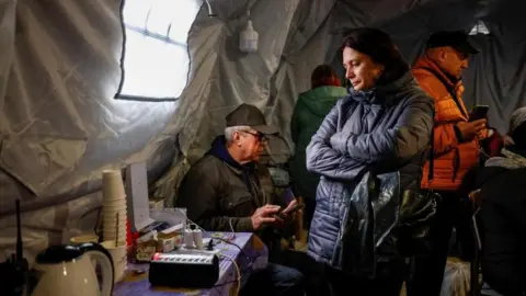 Reuters A woman wearing a dark-coloured winter coat stands next to a table with cups, a kettle and plug points on it in an "invincibility centre" -  where people can get an internet connection, charge their devices and warm up, amid a water and electricity supply outage in Chernihiv. A man sits on a chair behind the woman using his phone, while another is standing behind her also holding his phone. They are standing inside a large tent.
