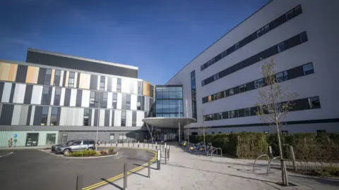 PA Media general view of the main entrance to the Royal Hospital for Children and Young People Edinburgh, a modern hospital building with sections of dark grey and light grey panelling and windows. There is a bike rack at the front and a road curves by the entrance