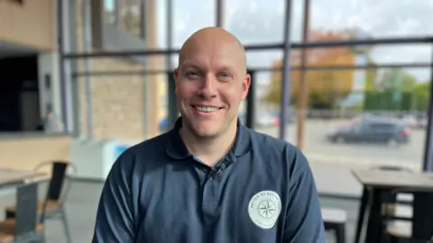 BBC Sam Coton wears a navy polo shirt with the 'Better Me, Better You' logo on it. He is sitting in the school theatre bar with floor to ceiling glass windows, you can see tables and chairs behind him. He is smiling at camera.