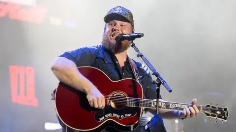 Getty Images Luke Combs performs during 2025 Austin City Limits Music Festival at Zilker Park. He has a brown beard is wearing a camo baseball cap, wearing a black short sleeve shirt and playing a guitar singing into a microphone.