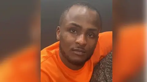 Met Police Sanchini Thompson looks at camera. He is wearing an orange T-shirt and has a short beard around his chin