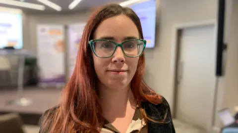 BBC A young woman with shoulder length auburn hair and wearing green framed glasses is looking at the camera. She is wearing a black jacket with a brown and cream blouse under it.