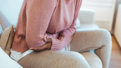 Getty Images A woman sitting on a cream sofa, with her arms folded around her stomach. the woman's face cannot be seen. She is wearing a pink jumper and beige leggings and is partly hunched over.