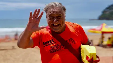 Supplied Mirek Craney, wearing a bright orange shirt with his name on it, waves at the camera. he is at the beach, a flotation device under his arm