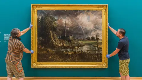 Alamy The large oil painting, Constable's Salisbury Cathedral from the Meadows, is being hung by two staff at the Tate Britain - its ornate gold frame contrasting against the teal coloured wall. In the foreground of the painting there is a horse drawn cart through the water meadows. Behind is Salisbury Cathedral and spire and in the distance some buildings, including Leaden Hall. A rainbow arcs through the cloudy sky towards the hall. 