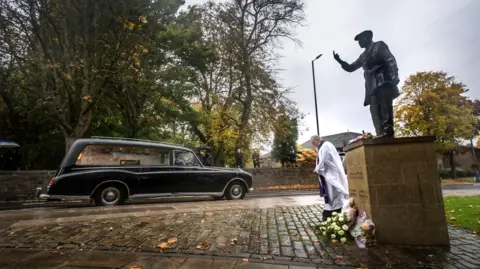 PA Media A hearse passing a statue with mourners lining the route