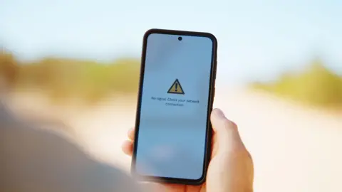 Getty Images A person holds a phone with a plain white background. On the screen is an error message stating there is no phone signal.