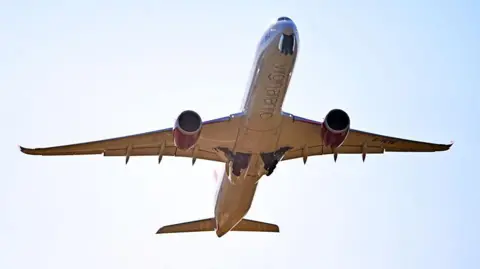 Getty Images A twin-engined plane seen from below, with Virgin Atlantic written on the white fuselage, with landing gear partially open