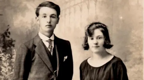 Family photo A black and white photograph showing a man with very dark hair wearing a suit, and a woman with dark hair down to her chin wearing a black blouse.