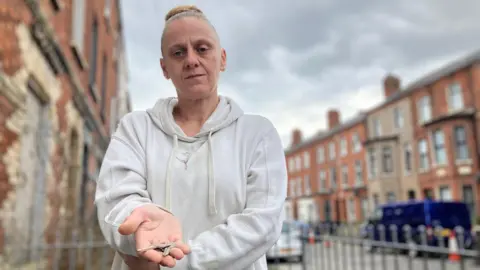 BBC A woman is standing in a small urban garden area with a long street behind her. It is lined with red brick houses and cars parked along the pavement. The woman has blonde hair tied up in a bun and is wearing a grey jumper. She looks directly at the camera while holding two loose keys in her hand. 