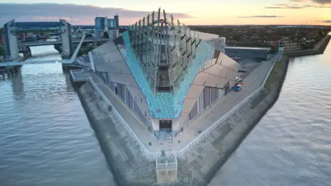 Getty Images A striking modern building in a triangular shape, with a glass front above blue and silver tiles, juts out on a promontory between two rivers under a yellow sky. To the left, bridges and a H-shaped tidal barrier can be seen straddling a river. To the right, houses can be seen stretching towards the horizon. 