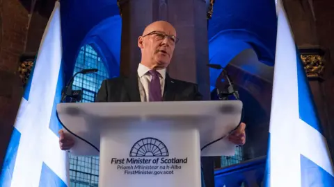 PA Media John Swinney, a bald man with glasses, dark suit and purple tie, gives a speech at a lectern saying "First Minister of Scotland". He is in the National Gallery of Scotland, a dark Gothic building, which has been lit in blue lights and two large Saltire flags flank him.