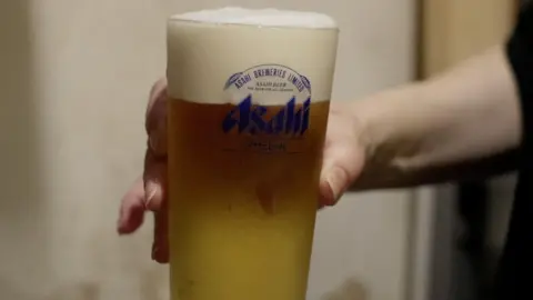 Reuters A person holds a large glass of beer in their right hand with Asahi Breweries written on the glass in blue letters.