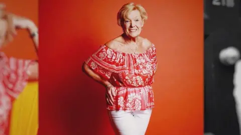 BBC Old woman with short blonde hair wearing a red top and white trousers posing for a photoshoot 