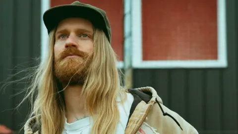 Jack Robinson Sam Ryder looks pensively into the distance, while standing in front of a wood cabin, whose windows and roof have been painted a distinctive shade of orange.