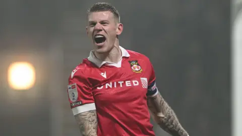 PA Media Footballer James mcClean pictured celebrating a goal. He is wearing the red strip of Wrexham FC, his arms are heavily tattooed