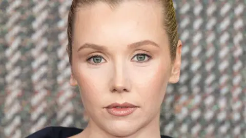 Getty Images A glamorous red carpet shot of actress Lauren Lyle is a close-up of her face, with a tweed-effect on the background behind her.
