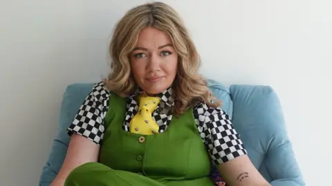 Serena Terry images Ms Terry is sitting on a light blue chair. She is looking at the camera with a confused expression. She is wearing a black and white checked t-shirt, yellow tie and green waistcoat and trousers. She has long blonde hair. It is a PR promo image.