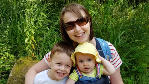 Family photo Nicola Wheatley is sitting on a  rock in a green space with her children on her lap. She and Oscar are smiling at the camera and  baby Ffion is wearing a yellow summer hat and dungarees. 