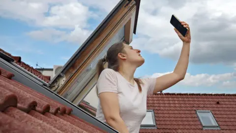Getty Images Young woman encounters signal problems with GPS or 5G on her smartphone while looking out of an open attic window, determined to restore connectivity.