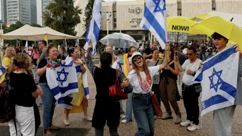 Reuters A group of people, with two women in front, linkinng hands and dancing, surrounded by others displaying Israeli flags, some with yellow umbrellas showing with 'NOW' printed on them  