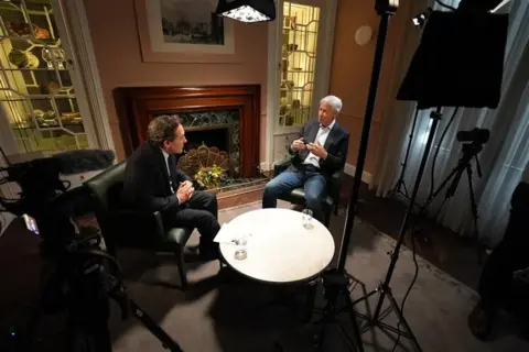BBC Business editor Simon Jack sits opposite Jamie Dimon as he interviews him near a fire place surrounded by TV cameras