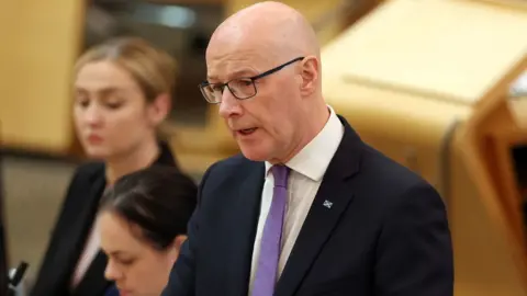 Getty Images John Swinney, who is bald with glasses, speaks in the Scottish Parliament chamber. He is wearing a dark suit, white shirt and purple tie 