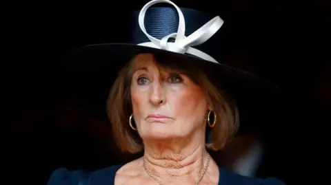Getty Images Annabel Goldsmith wearing a dark blue formal hat and dress. The hat has a cream-coloured bow. She is wearing gold hoop earrings. 
