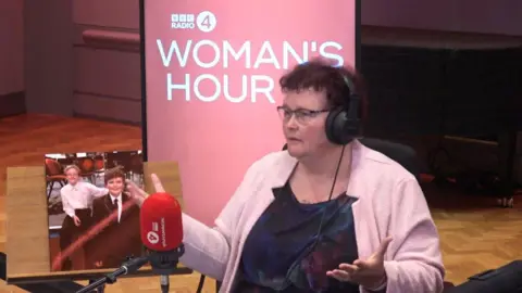 BBC A woman with brown curly hair wearing a dark blue dress and pink cardigan, during an interview for BBC Woman's Hour. She wears headphones and is in front of a microphone with the Woman's Hour logo behind her. There is a photo of two boys next to her.
