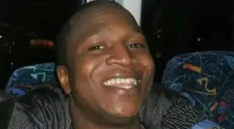 The Bayoh family A man with short black hair smiles for the camera. He is wearing a black top and appears to be sitting on a seat in a bus