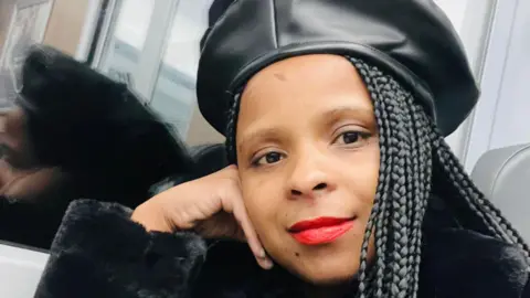 Facebook Naso Isaacs loking straight at the camera. She is wearing a black leather beret and has long black hair. Her head is propped up on her right hand. She has red lips.