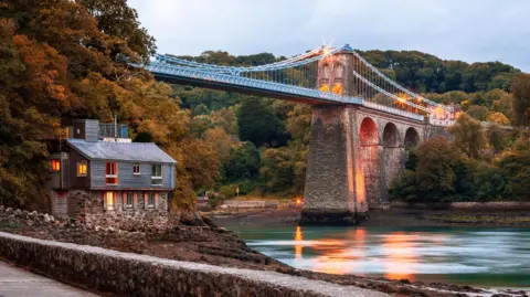 Getty Images Bridge seen from one side and below, along a path with view of the river below and a house to the left. The bridge is lit in warm yellow light 