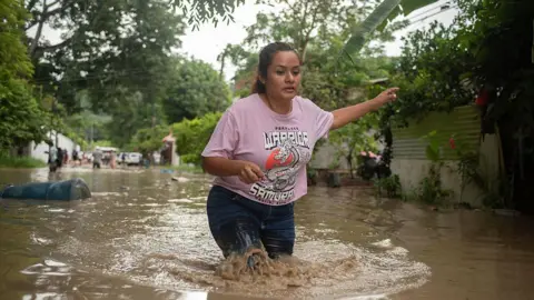 Getty Images A woman in a pink t-shirt and jeans wades through a flooded street, with muddy water up to her knees