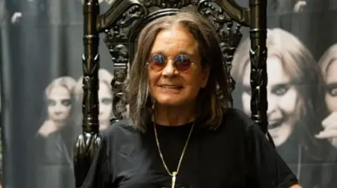 Getty Images Ozzy Osbourne sits on a dark wood, high-backed chair with ornate designs on it. He is wearing round sunglasses, and is smiling, while also wearing a black T-shirt and a silver pendant chain. Behind him there are images of him on canvas with different facial portraits of the musician.