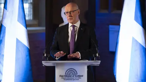 PA Media John Swinney looking slightly off camera. He is standing at a podium between two Scotland flags. His hands are raised slightly above the top of the podium. He is wearing a dark suit over a white shirt with a purple tie.