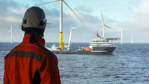 Getty Images A person wearing orange overalls and a hard hat looks out over the North Sea. There is a ship next to a large wind pylon in the centre of the shot, with other pylons in the background. 