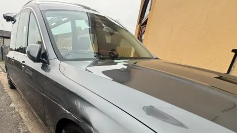 Martin Vowles Funeral Directors A grey Mercedes hearse parked beside a yellow wall. It has a long scratch down the side above the wheel. 