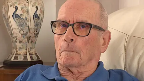 BBC Des - an older-looking man looks at the camera. He is wearing black-rimmed glasses and a blue polo shirt. It looks like he's sat on a sofa with a vase behind.