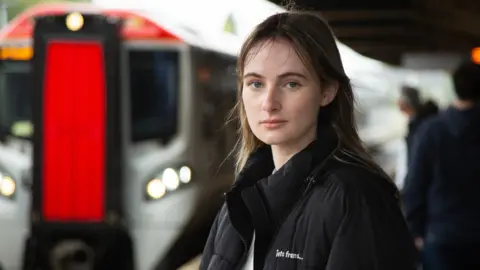 BBC A woman with long dark hair wearing a black coat and white T-shirt stands in front of a train with a red front door at a station