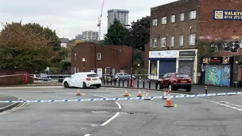 BBC/Chloe Aslett This image captures a cordoned-off street. A white car is covered with a protective sheet, suggesting it may be central to the incident. Nearby, a red car is parked, and the area is marked off with police tape and traffic cones to restrict access. The surroundings include buildings with signage and graffiti art, and in the distance, a crane and tall structures indicate a city environment.